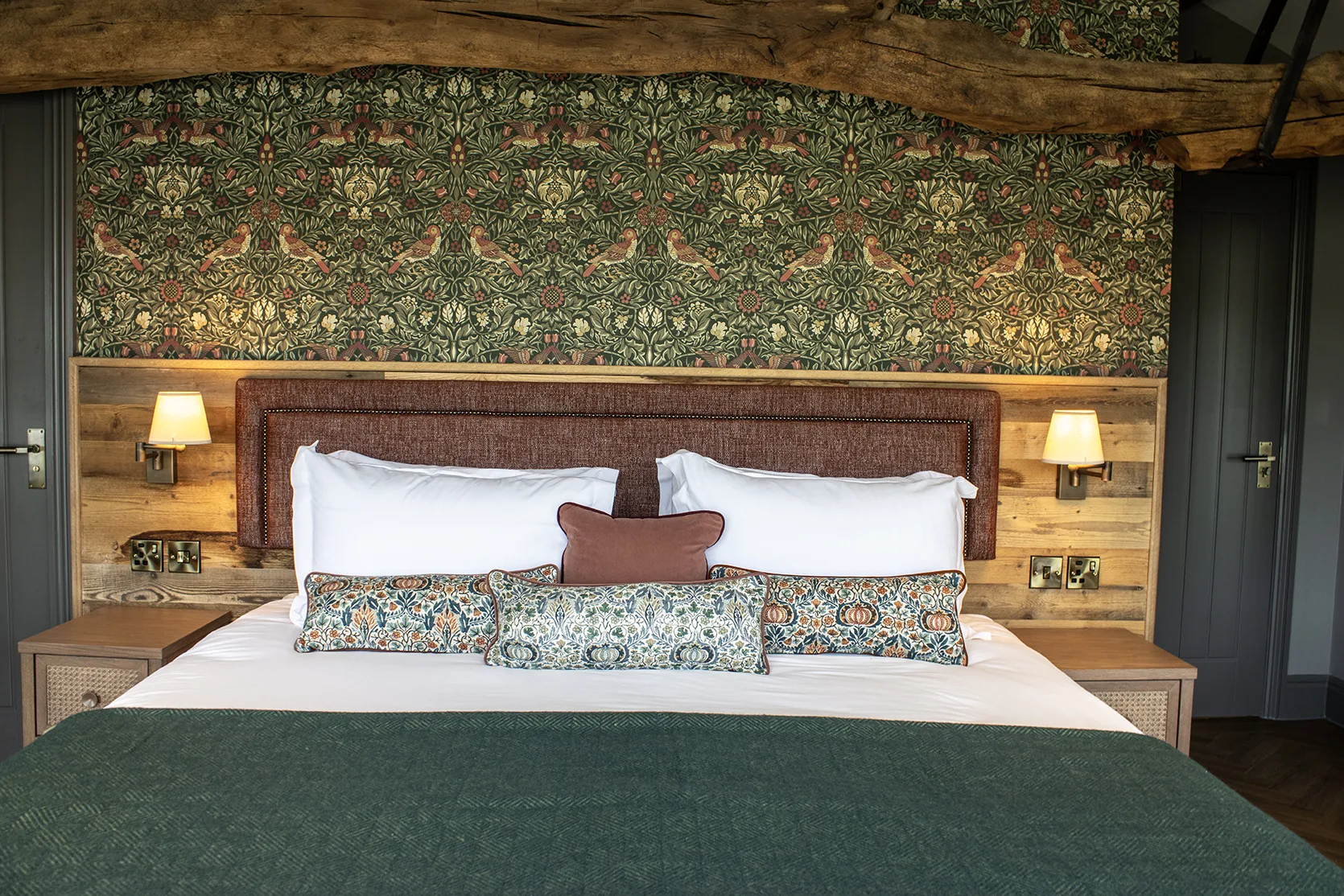 King-size bed with crisp white bedding, patterned cushions and a green wool throw, set against a dark green bird and foliage wallpaper, with a rustic wooden headboard wall, wall lamps and an exposed oak beam above.